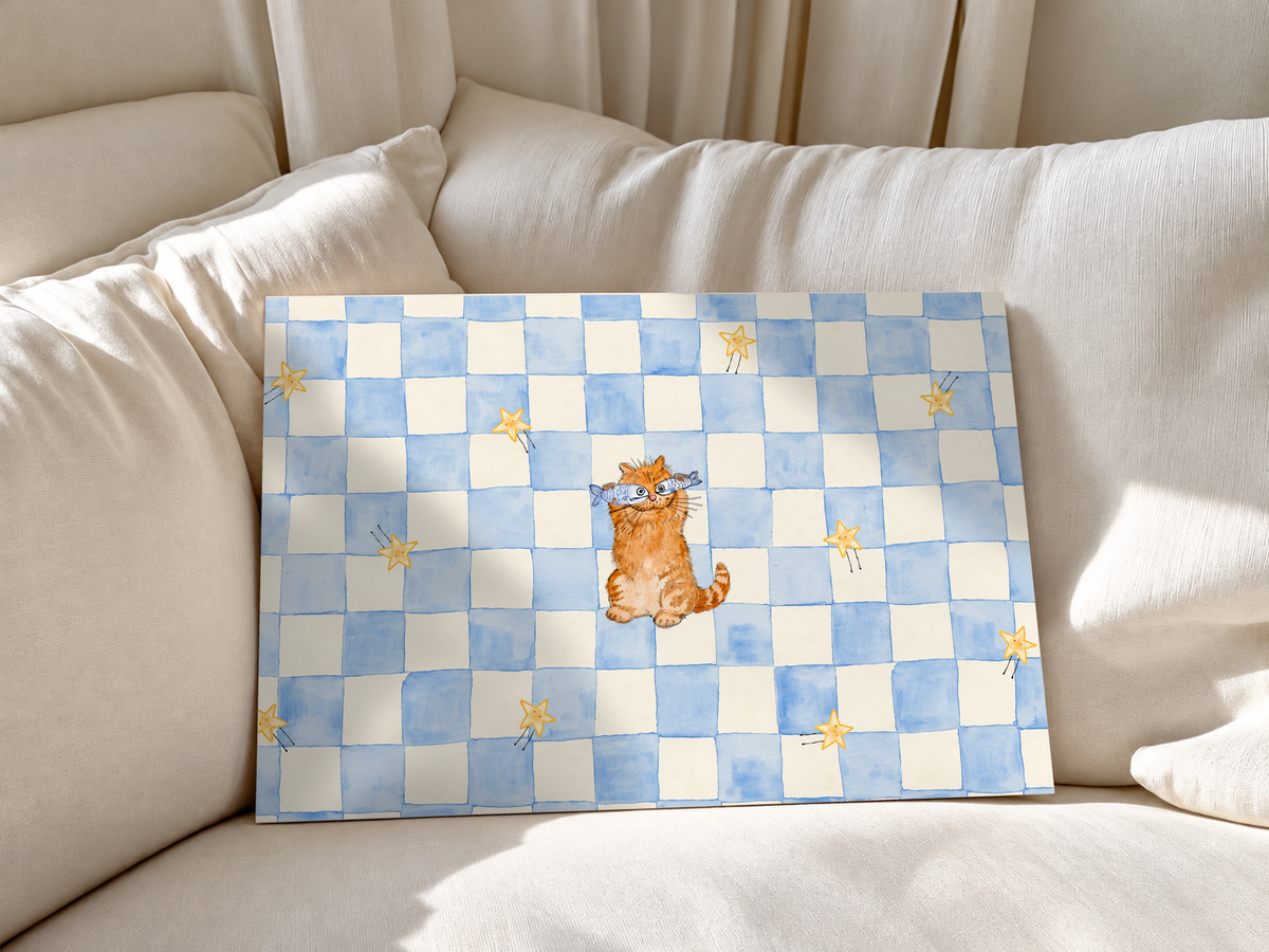 Un pequeño gato naranja con antifaz azul claro se sienta entre estrellas doradas sobre un tablero azul y crema. Póster sin marco apoyado en cojines blancos de sofá.