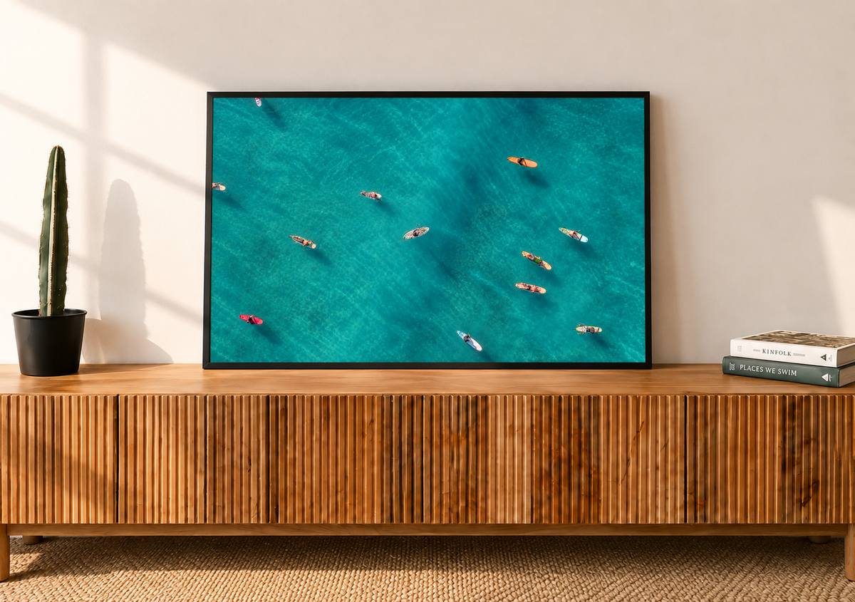 Vista dall’alto di surfisti seduti o sdraiati su tavole arancioni, rosse e chiare sparse sull’acqua turchese. Cornice nera su una credenza in legno a listelli, accanto a un cactus in vaso e a libri.
