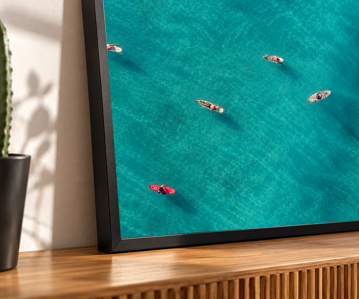 Vista dall’alto di surfisti seduti o sdraiati su tavole arancioni, rosse e chiare sparse sull’acqua turchese. Dettaglio di una cornice nera appoggiata su una credenza in legno, con un cactus a sinistra.