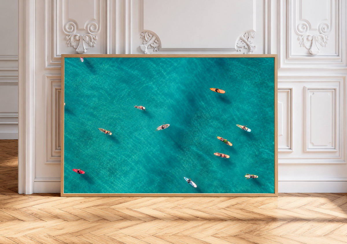 Vista dall’alto di surfisti seduti o sdraiati su tavole arancioni, rosse e chiare sparse sull’acqua turchese. Cornice dall’aspetto rovere appoggiata a una parete bianca con modanature e pavimento a spina di pesce.
