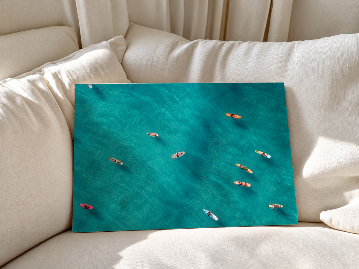 Vista dall’alto di surfisti seduti o sdraiati su tavole arancioni, rosse e chiare sparse sull’acqua turchese. Poster senza cornice appoggiato a morbidi cuscini bianchi del divano.