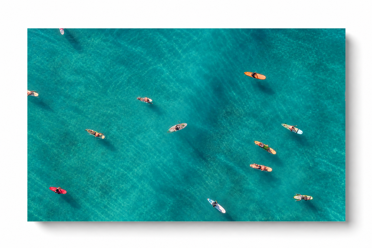 Vista dall’alto di surfisti seduti o sdraiati su tavole arancioni, rosse e chiare sparse sull’acqua turchese. Poster orizzontale senza cornice su fondo bianco con una morbida ombra.
