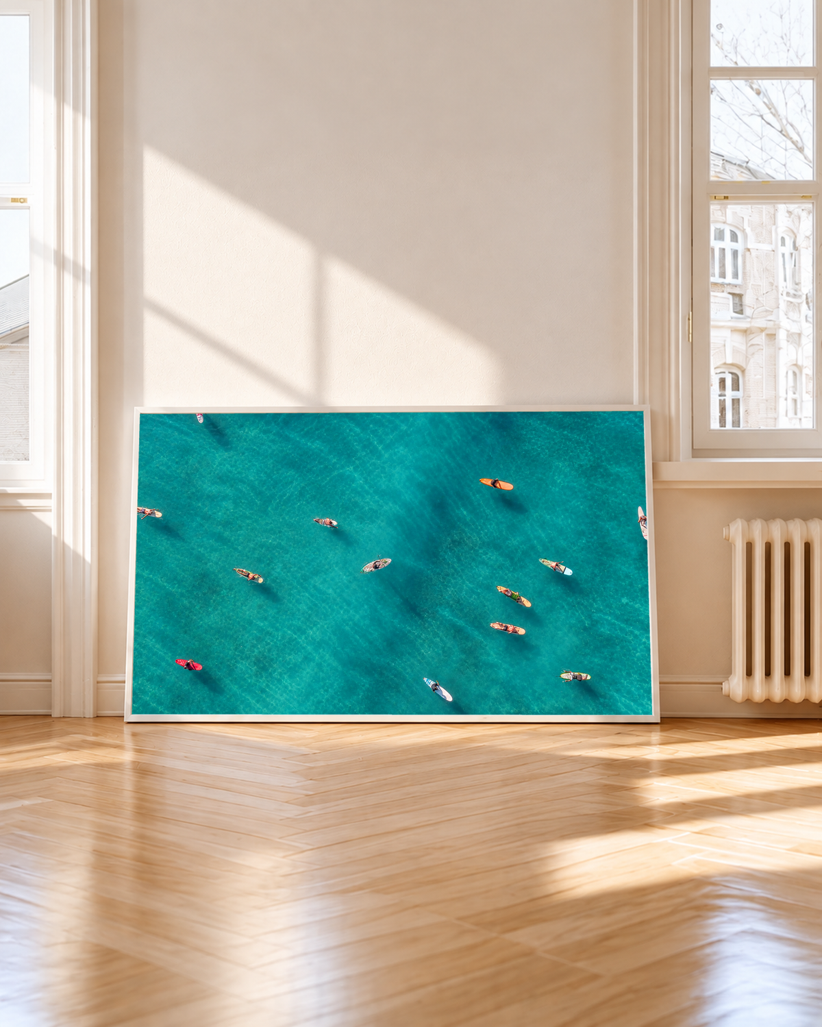 Vista dall’alto di surfisti seduti o sdraiati su tavole arancioni, rosse e chiare sparse sull’acqua turchese. Cornice bianca accanto a una finestra alta, con sole e ombre sul pavimento in legno.