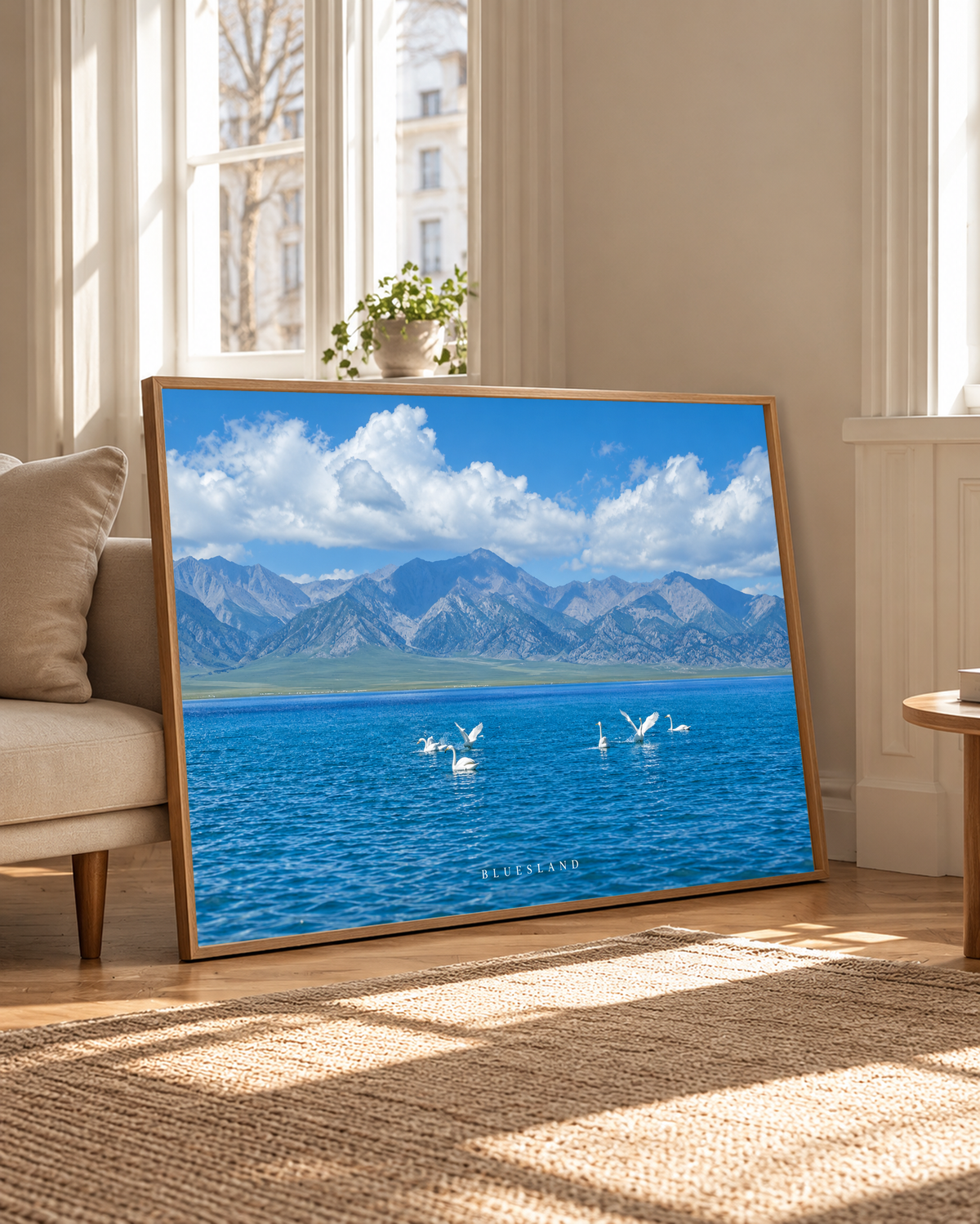 Weiße langhalsige Wasservögel schwimmen und breiten ihre Flügel auf tiefblauem Wasser vor einer graublauen Bergkette und hellen Wolken aus. Eichenfarbener Rahmen neben einem hellen Polstersessel und hohen Fenstern im warmen Tageslicht.