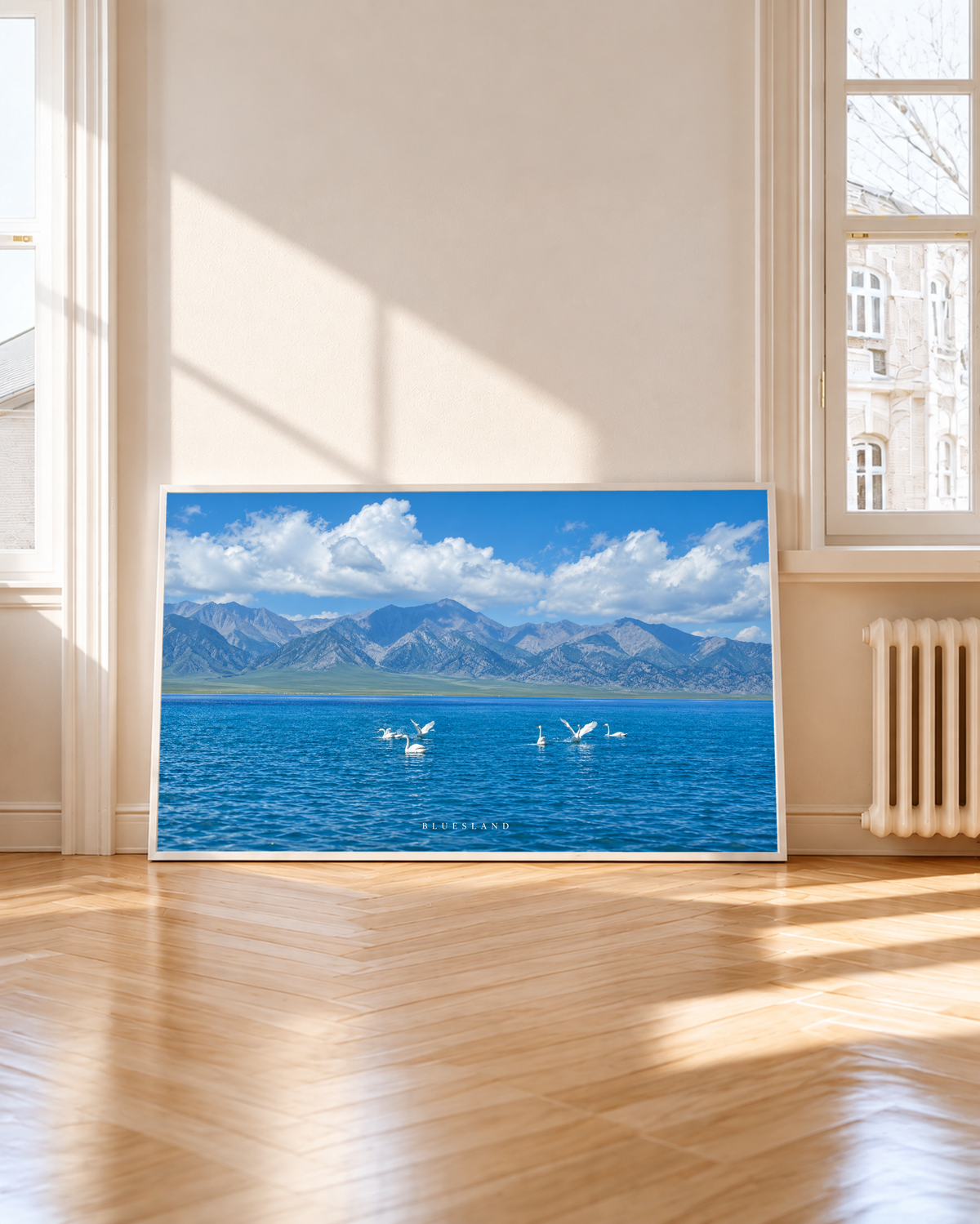 Weiße langhalsige Wasservögel schwimmen und breiten ihre Flügel auf tiefblauem Wasser vor einer graublauen Bergkette und hellen Wolken aus. Weißer Rahmen neben einem hohen Fenster mit Sonnenlicht und Schatten auf dem Holzboden.