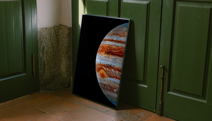 Close-up Jupiter artwork with orange-brown and blue-white cloud bands and the Great Red Spot on black, styled in a black frame against dark green paneled doors above orange floor tiles