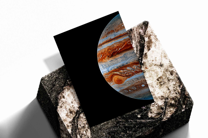 Close-up Jupiter artwork with orange-brown and blue-white cloud bands and the Great Red Spot on black, shown as an unframed print tilted on a dark stone display against a light background
