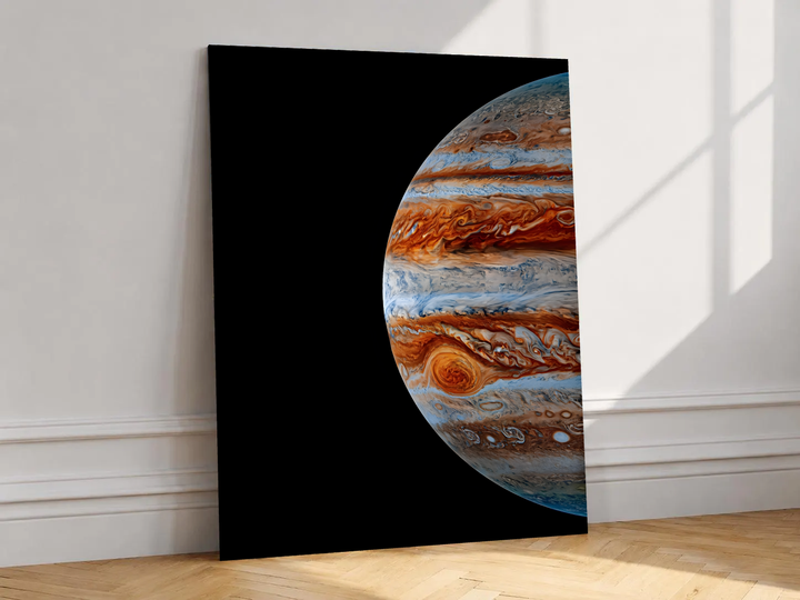Close-up Jupiter artwork with orange-brown and blue-white cloud bands and the Great Red Spot on black, shown unframed on a white wall above parquet flooring with a diagonal sun shadow