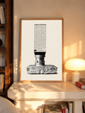 A monochrome Leica camera beneath a tall column of advertising typography in an oak frame above a pale wooden sideboard beside a glowing lamp