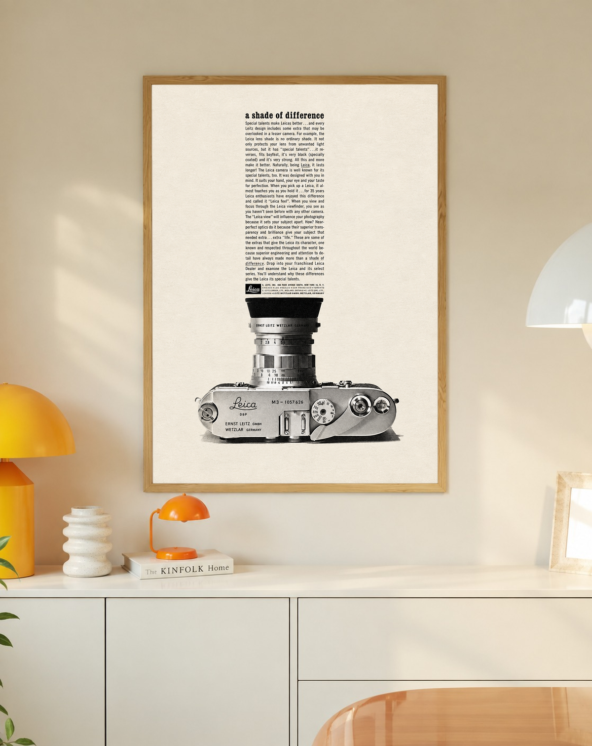 A monochrome Leica camera beneath a tall column of advertising typography in a slim oak frame on a cream wall above a white cabinet