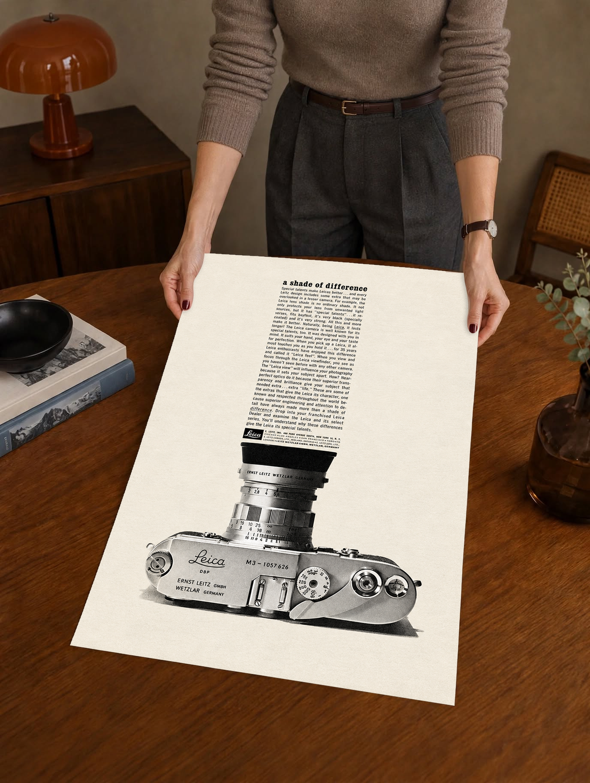 A monochrome Leica camera beneath a tall column of advertising typography as a large unframed poster held above a wooden desk