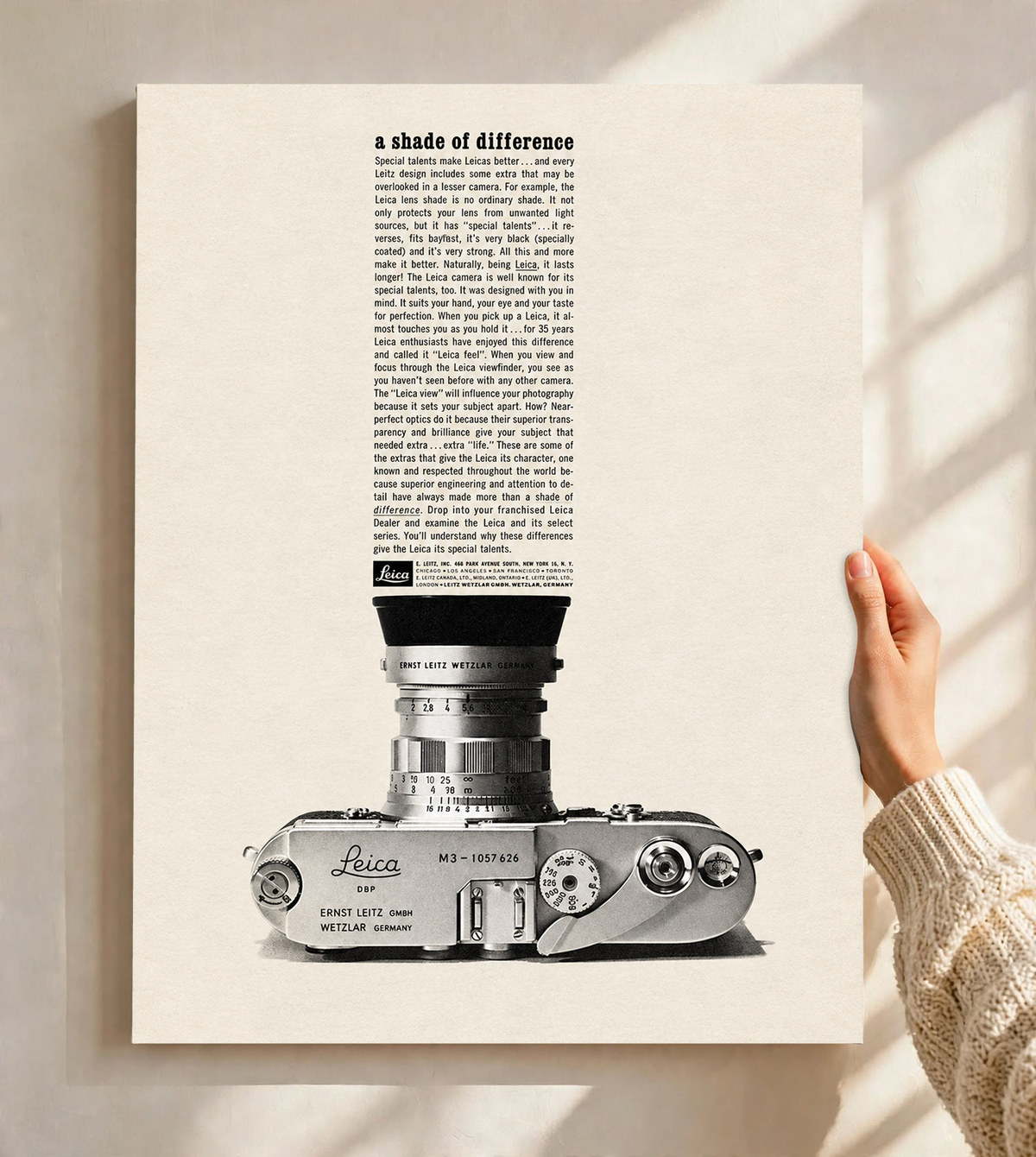 A monochrome Leica camera beneath a tall column of advertising typography as an unframed poster held against a pale sunlit wall
