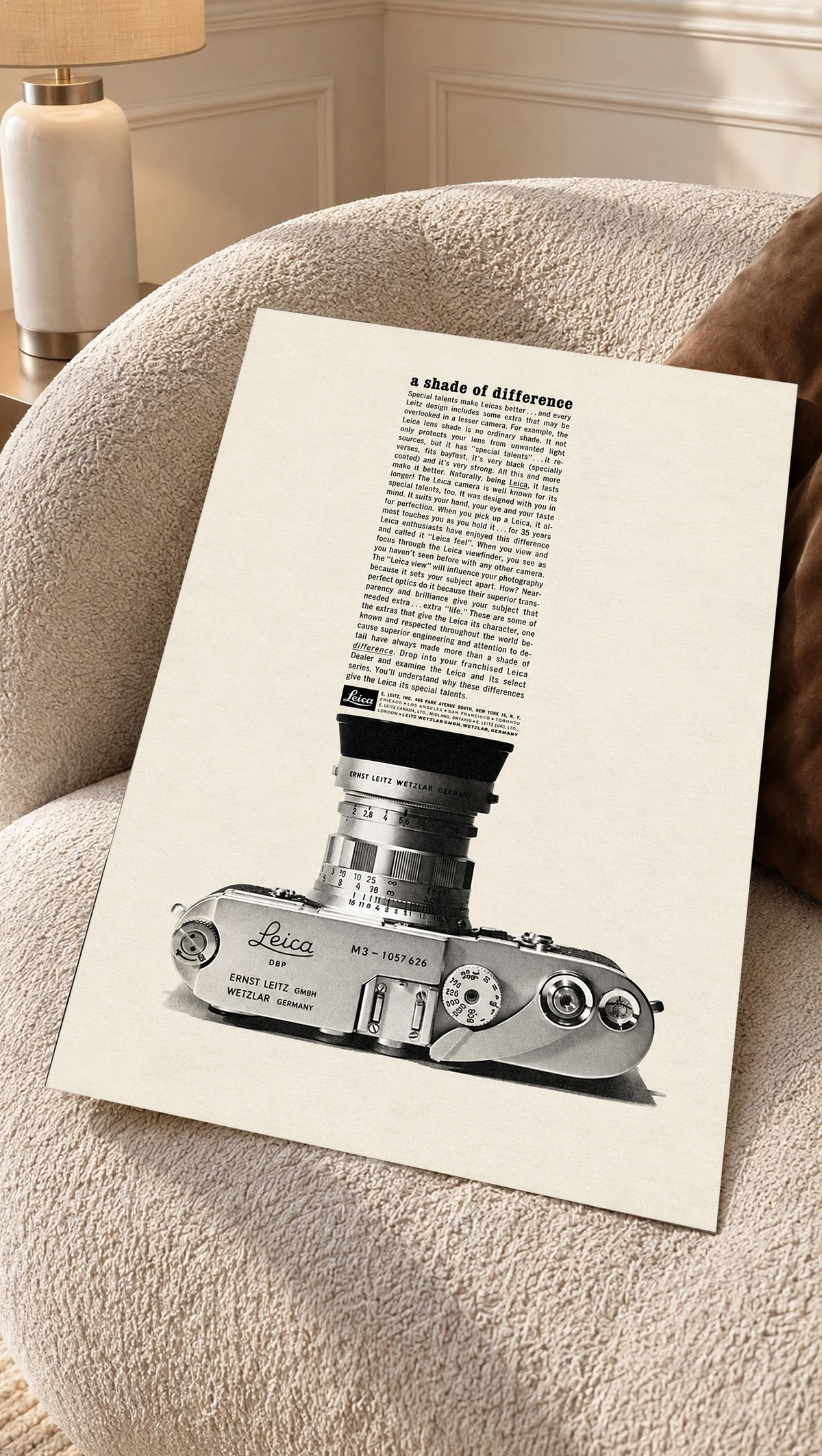 A monochrome Leica camera beneath a tall column of advertising typography as an unframed poster resting on a cream woven chair