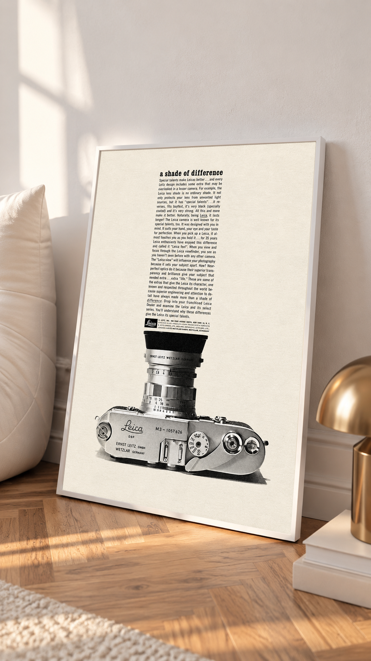 A monochrome Leica camera beneath a tall column of advertising typography in a white frame on a sunlit floor beside a white cabinet