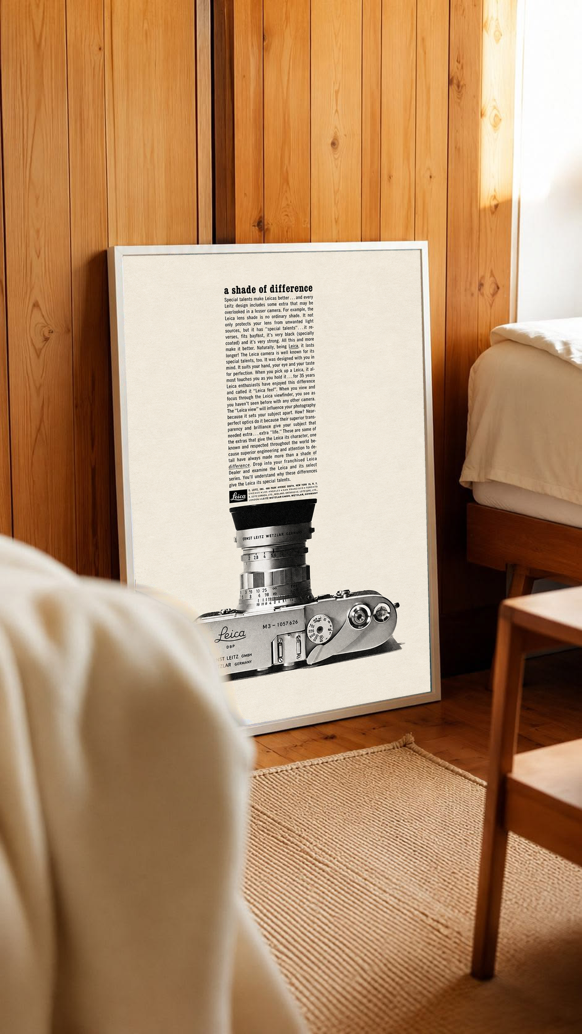 A monochrome Leica camera beneath a tall column of advertising typography in a white frame leaning against warm wooden wall panels