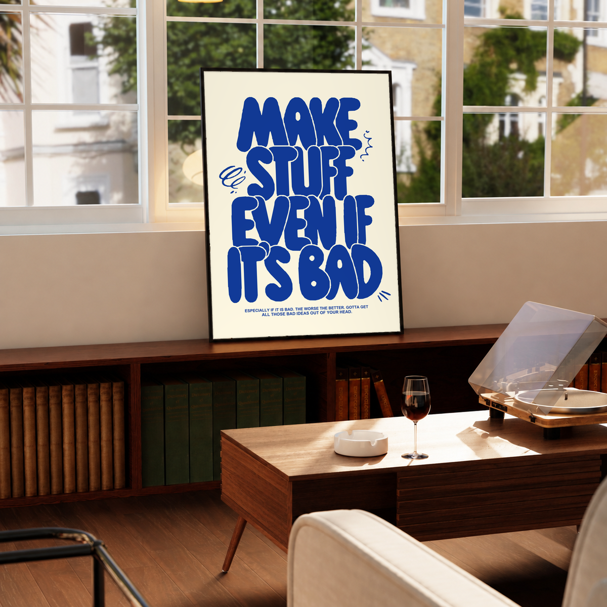 Framed cream poster with bold blue “MAKE STUFF EVEN IF IT'S BAD” on a tabletop.