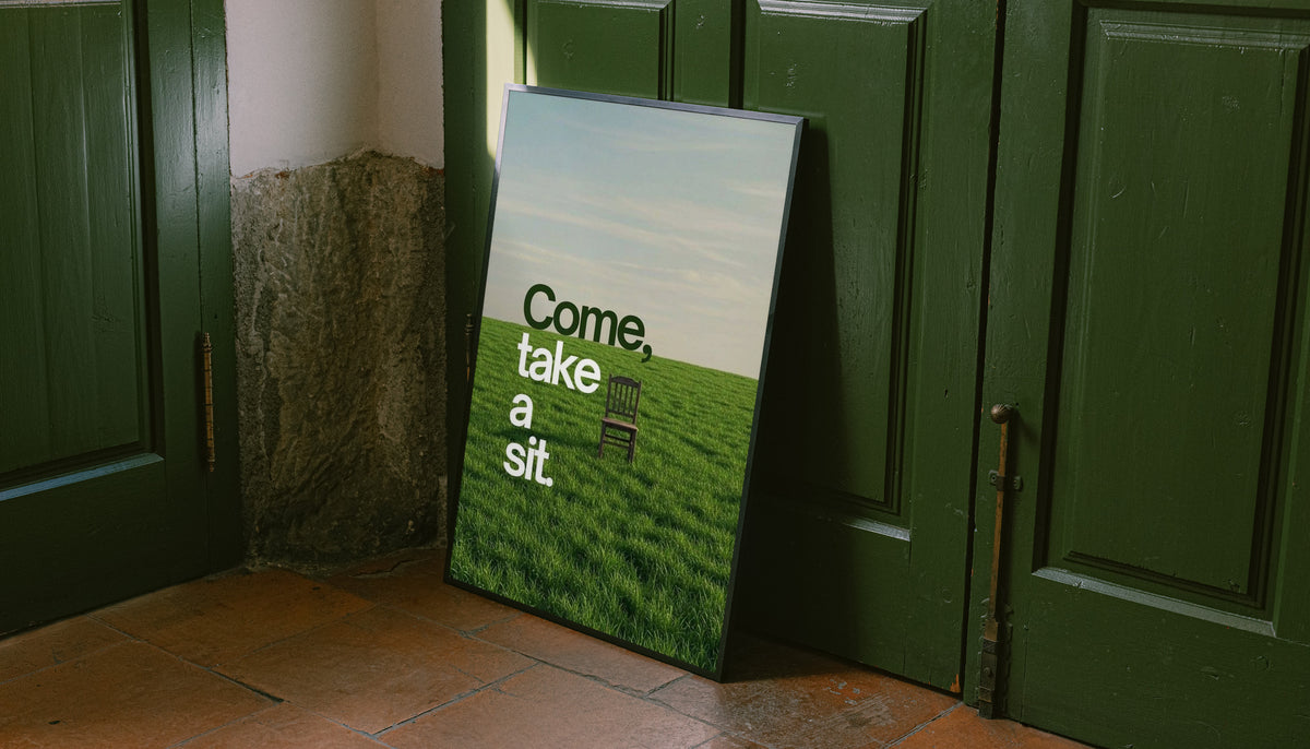 Landscape quote poster leaning near door, with green grass and text "Come, take a sit."