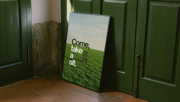 Landscape quote poster leaning near door, with green grass and text "Come, take a sit."