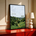 Green valley and winding path beneath Meet You Like the Wind lettering; black framed print on a red cabinet