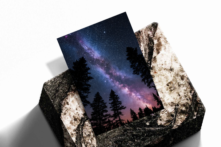 Violet-pink Milky Way band across a dark blue starry sky above black conifer silhouettes, shown as an unframed print tilted on a dark stone display against a light background