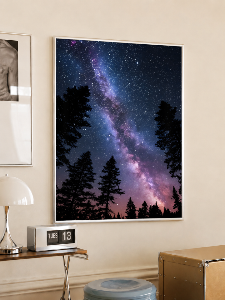 Violet-pink Milky Way band across a dark blue starry sky above black conifer silhouettes, styled in a light frame on a cream wall beside a white dome lamp and glass side table