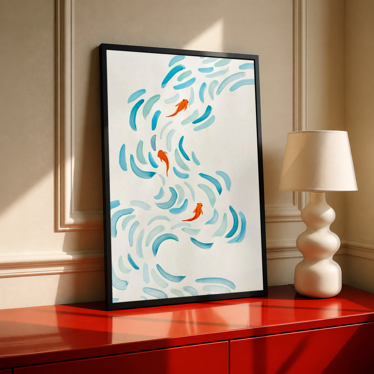 Small orange koi swimming among curved aqua and blue brushstrokes on cream in a black frame on a red console beside a sculptural cream lamp