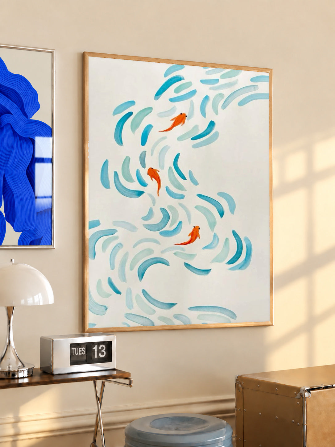 Small orange koi swimming among curved aqua and blue brushstrokes on cream in a light wood frame on a sunlit office wall beside a desk