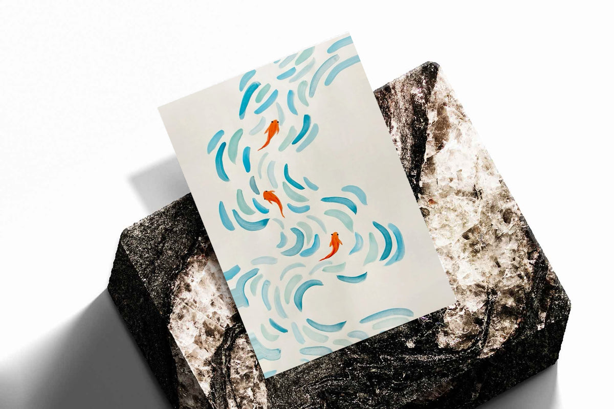 Small orange koi swimming among curved aqua and blue brushstrokes on cream as an unframed print resting at an angle on a dark stone display