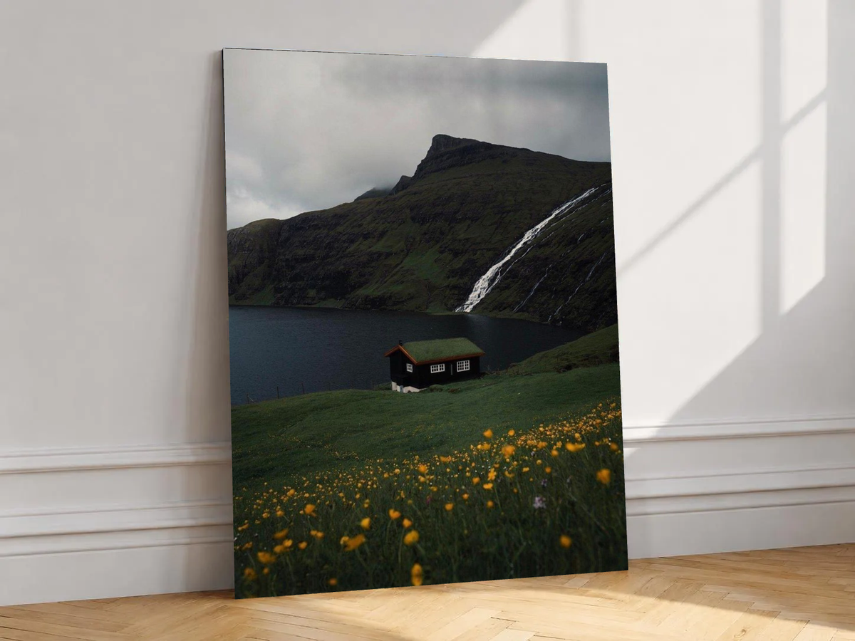Canvas artwork depicting lakeside cottage beneath a waterfall, leaning against a white wall in angled sunlight.