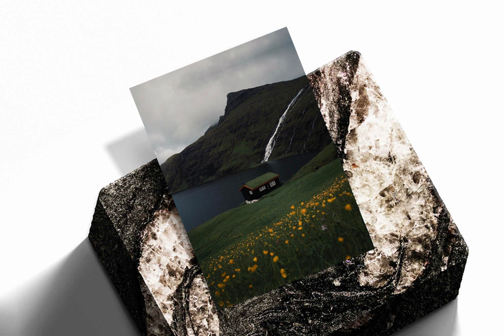 Unframed artwork depicting lakeside cottage beneath a waterfall, presented diagonally on rough stone.