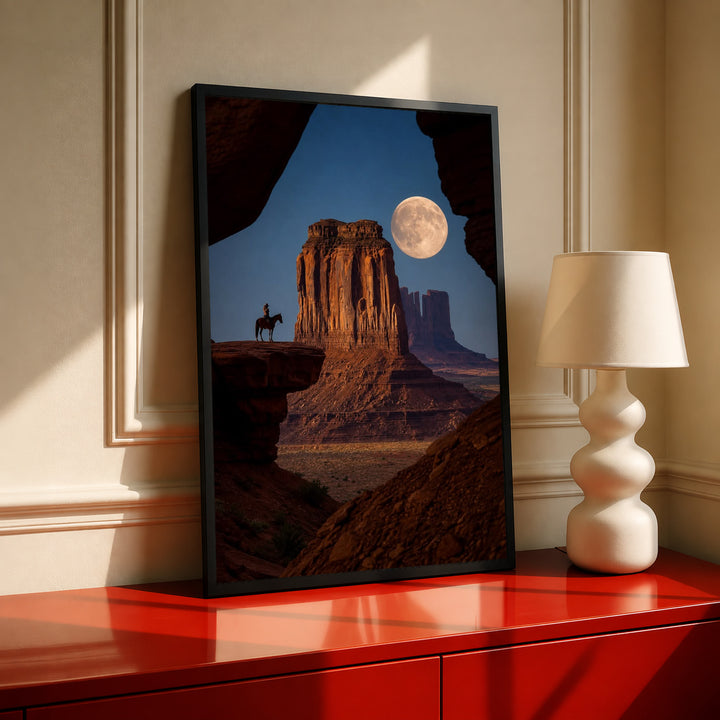 A horseback rider silhouetted beside a sandstone desert butte beneath a full moon in a black frame on a red console beside a sculptural cream lamp