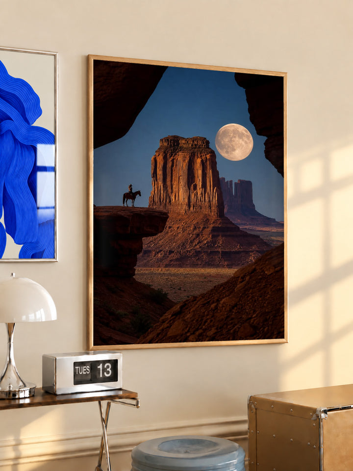 A horseback rider silhouetted beside a sandstone desert butte beneath a full moon in a light wood frame on a sunlit office wall beside a desk