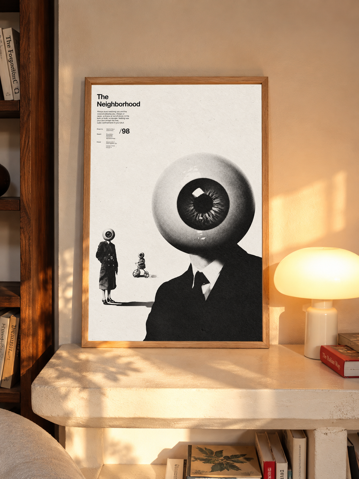 A suited eyeball-headed figure with smaller surreal figures on cream in an oak frame above a pale wooden sideboard beside a glowing lamp