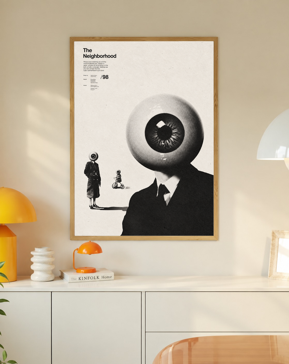 A suited eyeball-headed figure with smaller surreal figures on cream in a slim oak frame on a cream wall above a white cabinet