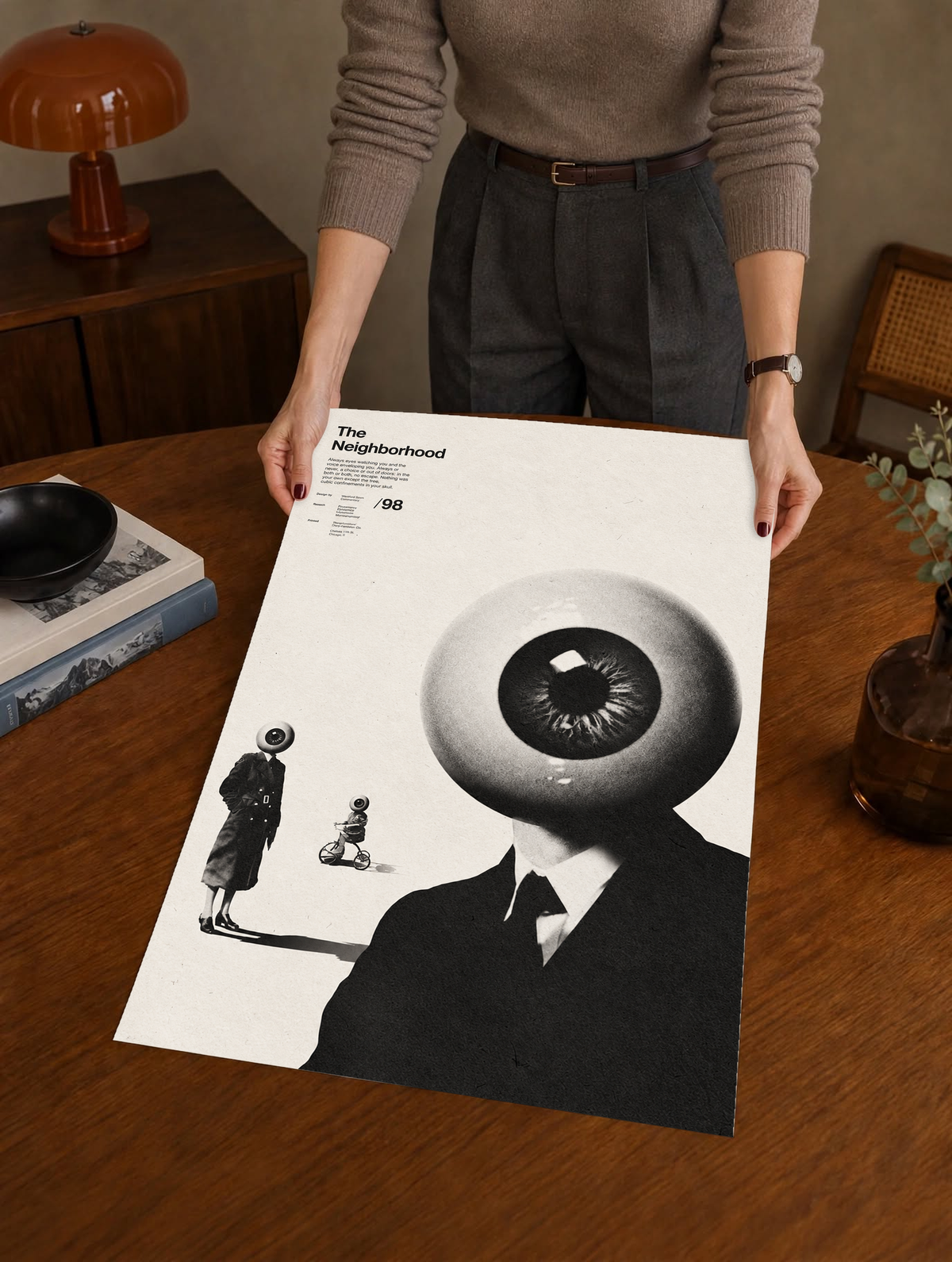 A suited eyeball-headed figure with smaller surreal figures on cream as a large unframed poster held above a wooden desk