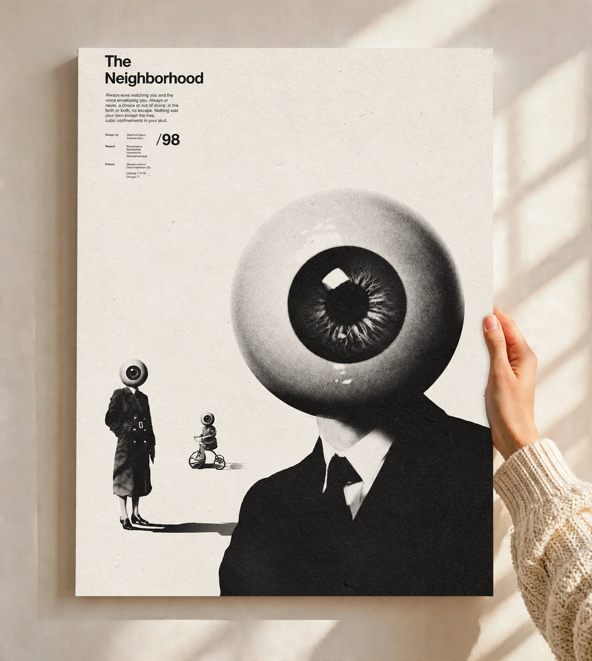 A suited eyeball-headed figure with smaller surreal figures on cream as an unframed poster held against a pale sunlit wall