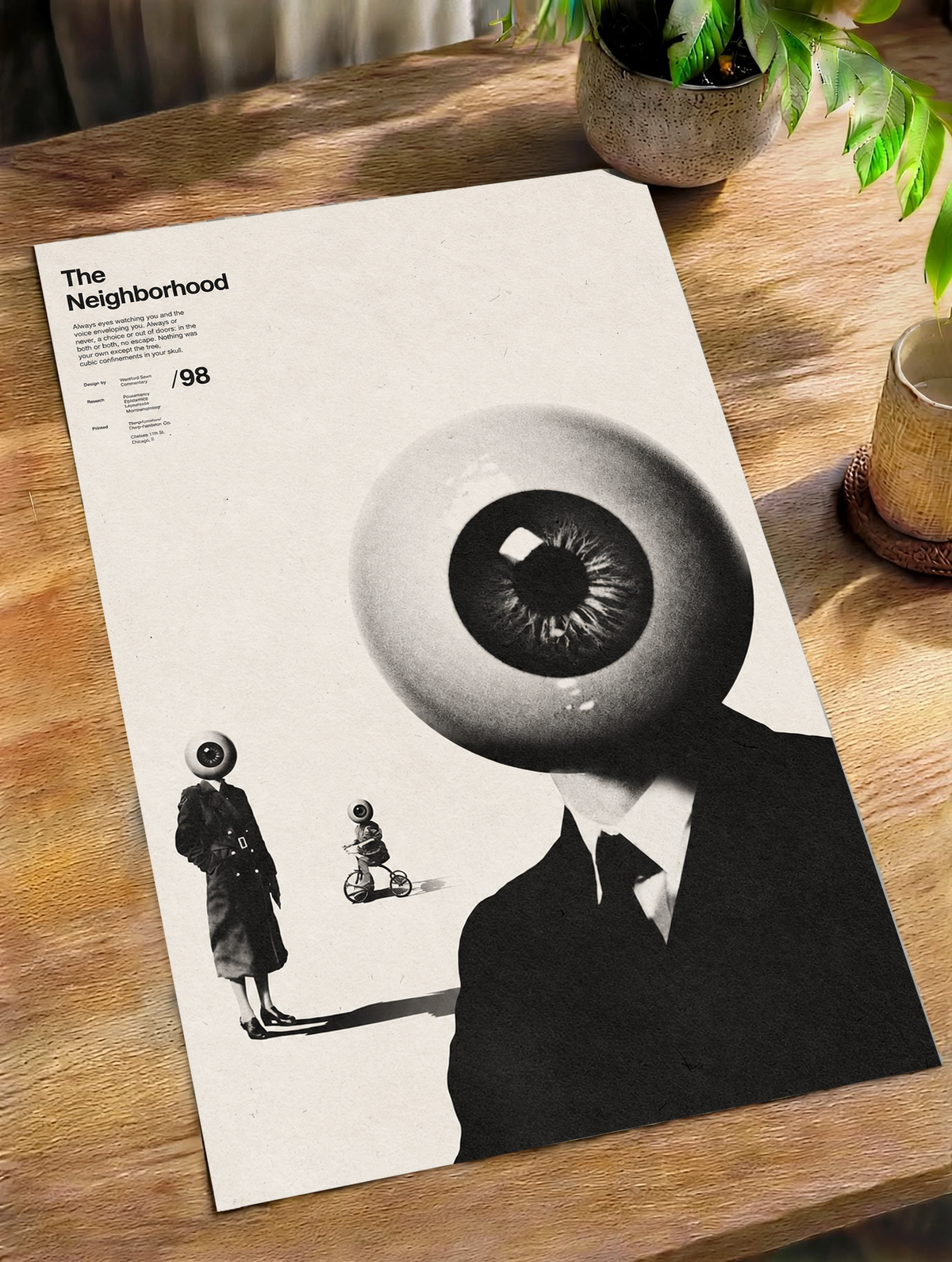 A suited eyeball-headed figure with smaller surreal figures on cream as an unframed poster lying on a wooden table beside a plant