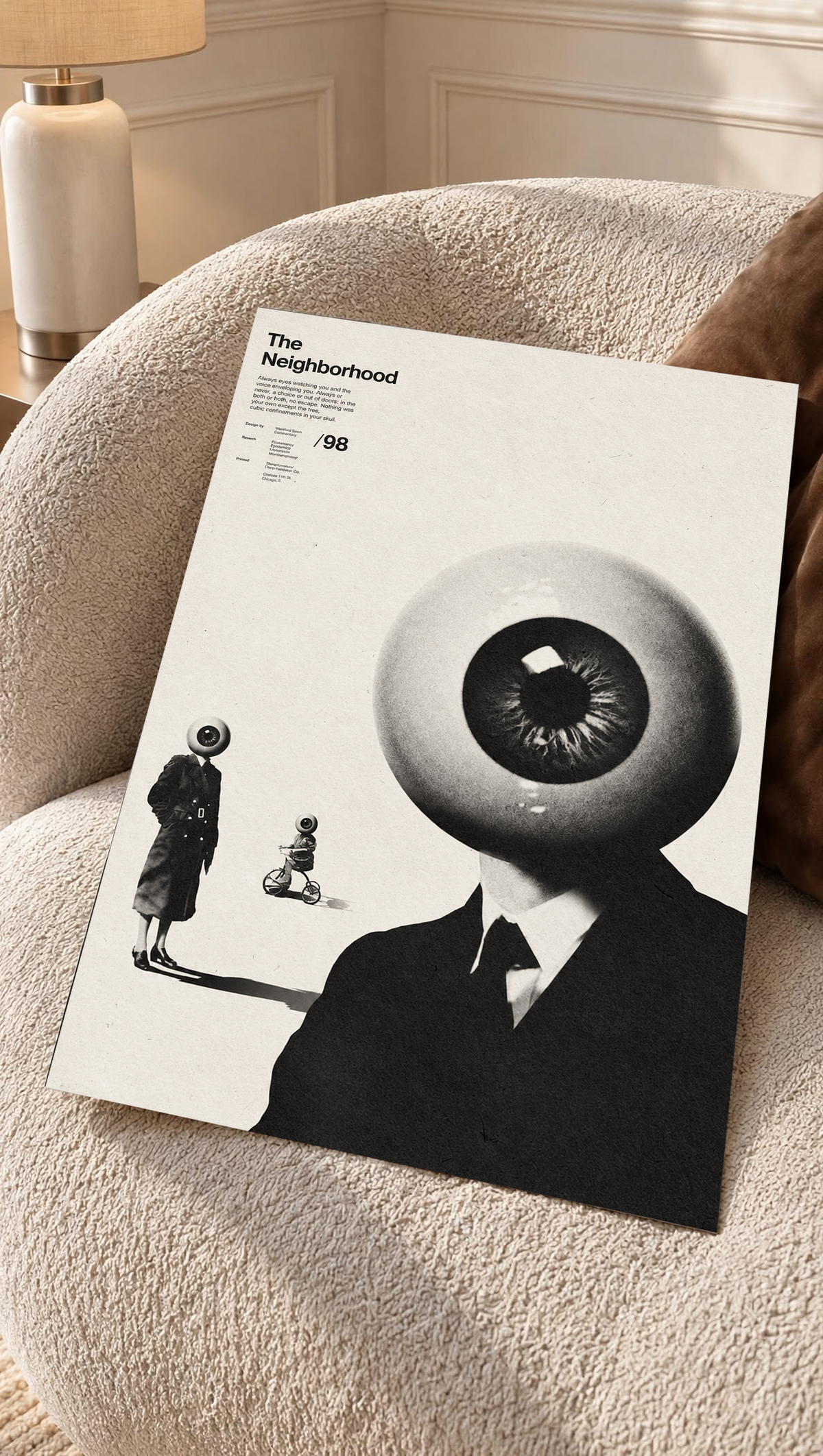 A suited eyeball-headed figure with smaller surreal figures on cream as an unframed poster resting on a cream woven chair