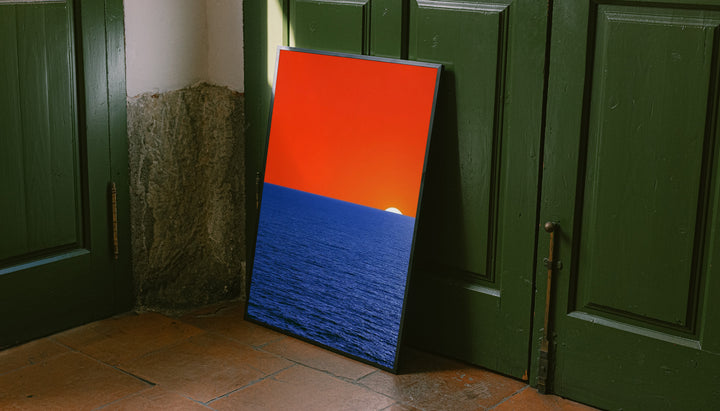 Tall framed coastal sunset wall art with orange sky, blue ocean, and white sun