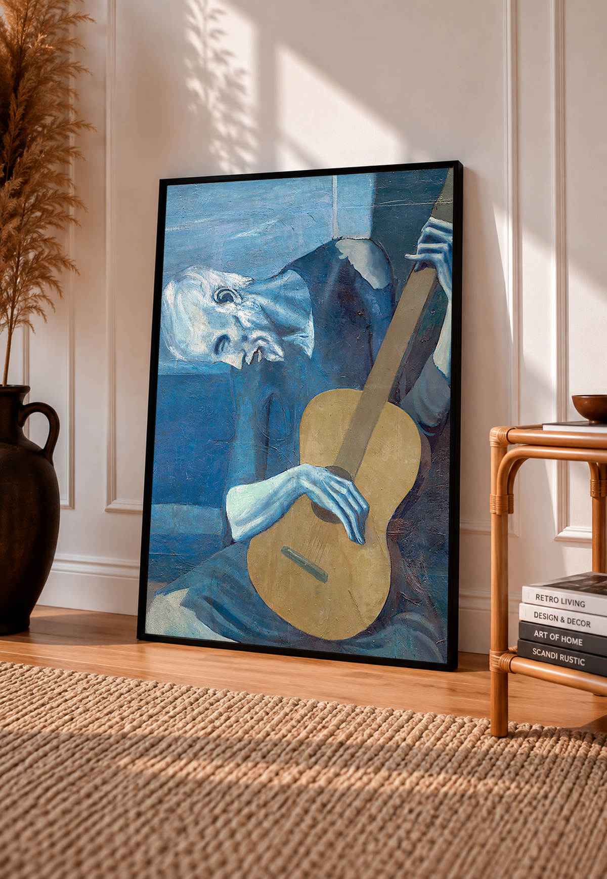 Picasso The Old Guitarist cropped portrait with a bowed blue figure and ochre guitar in a black frame leaning on a pale panelled wall beside a rattan chair