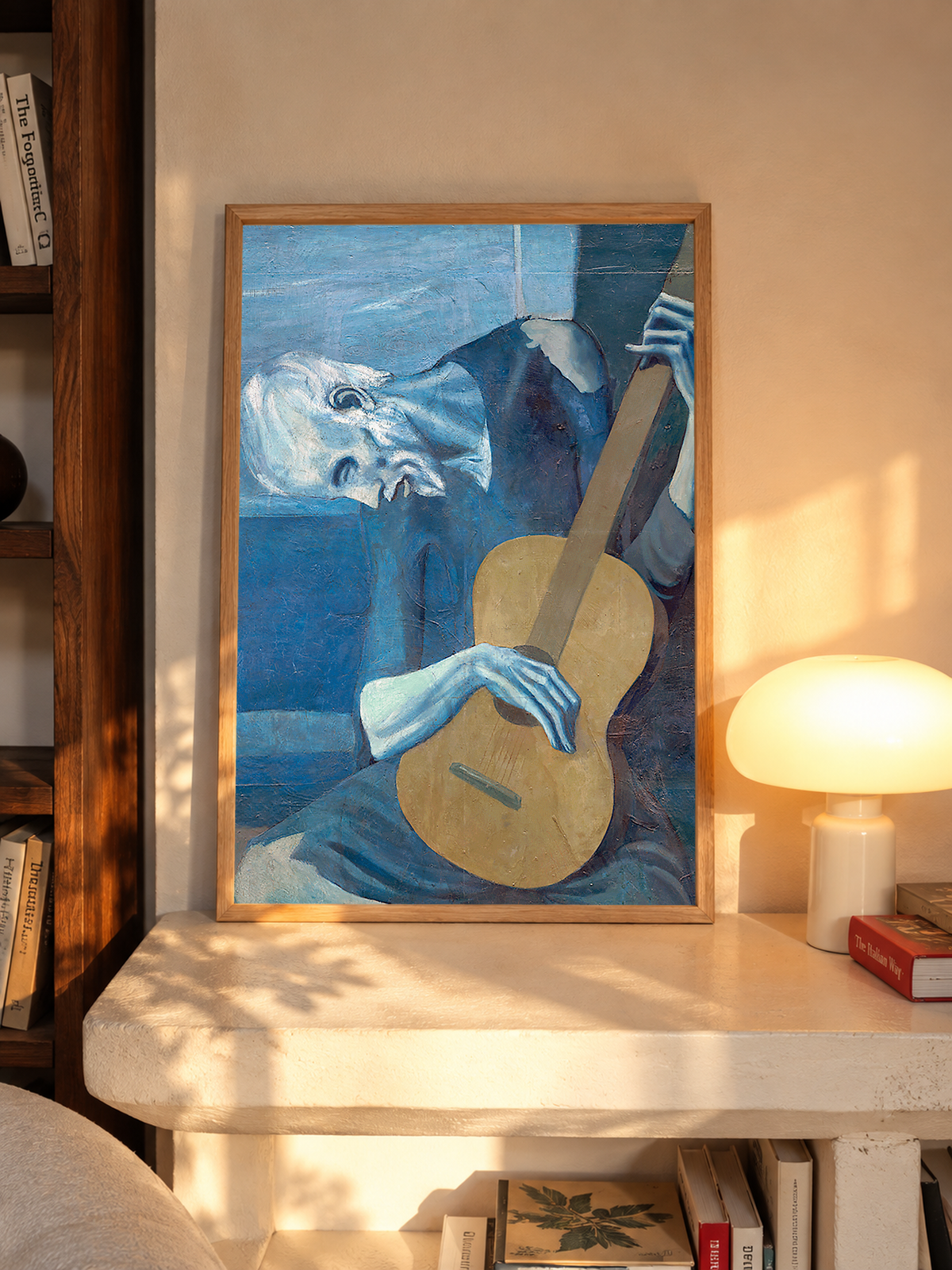 Picasso The Old Guitarist cropped portrait with a bowed blue figure and ochre guitar in an oak frame on a sunlit console beside books and a cream lamp