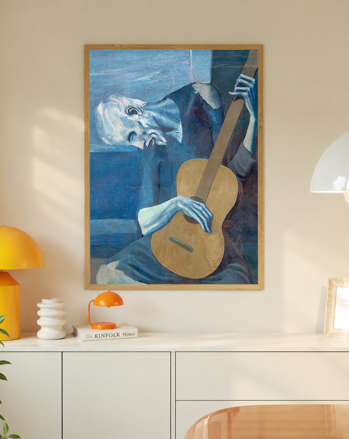 Picasso The Old Guitarist cropped portrait with a bowed blue figure and ochre guitar in an oak frame on a wall above a white cabinet beside a yellow lamp
