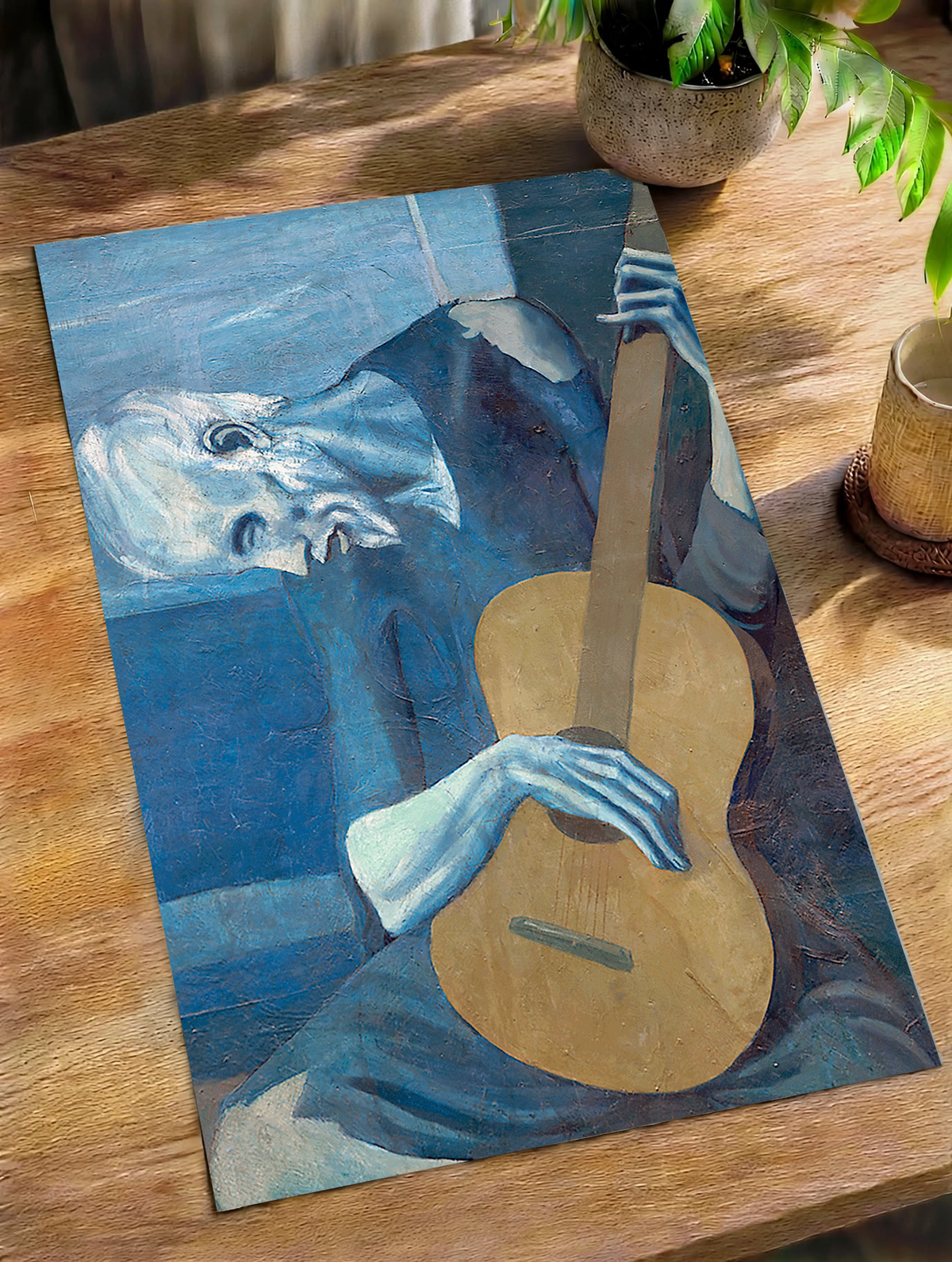 Picasso The Old Guitarist cropped portrait with a bowed blue figure and ochre guitar as an unframed print on a wooden table beside a plant