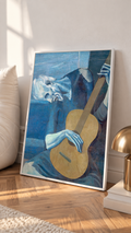 Picasso The Old Guitarist cropped portrait with a bowed blue figure and ochre guitar in a white frame leaning on a sunlit floor beside a brass lamp