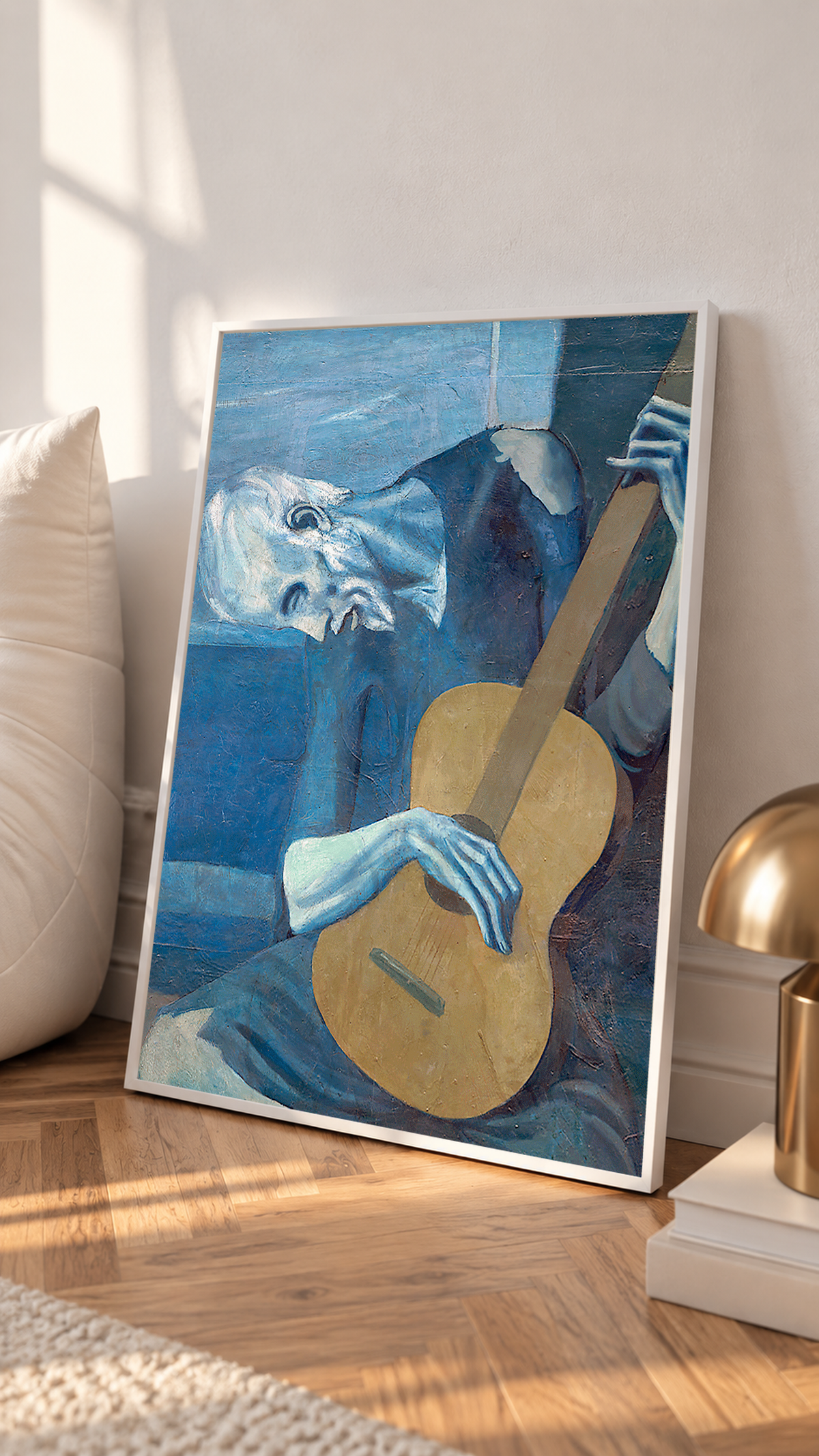 Picasso The Old Guitarist cropped portrait with a bowed blue figure and ochre guitar in a white frame leaning on a sunlit floor beside a brass lamp
