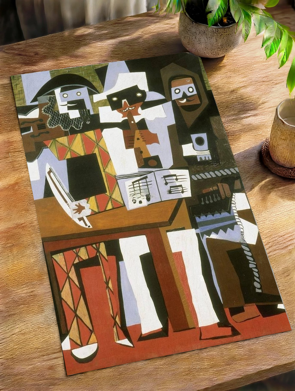 Picasso Three Musicians with angular masked figures, red diamonds and an accordion as an unframed print on a wooden table beside a plant
