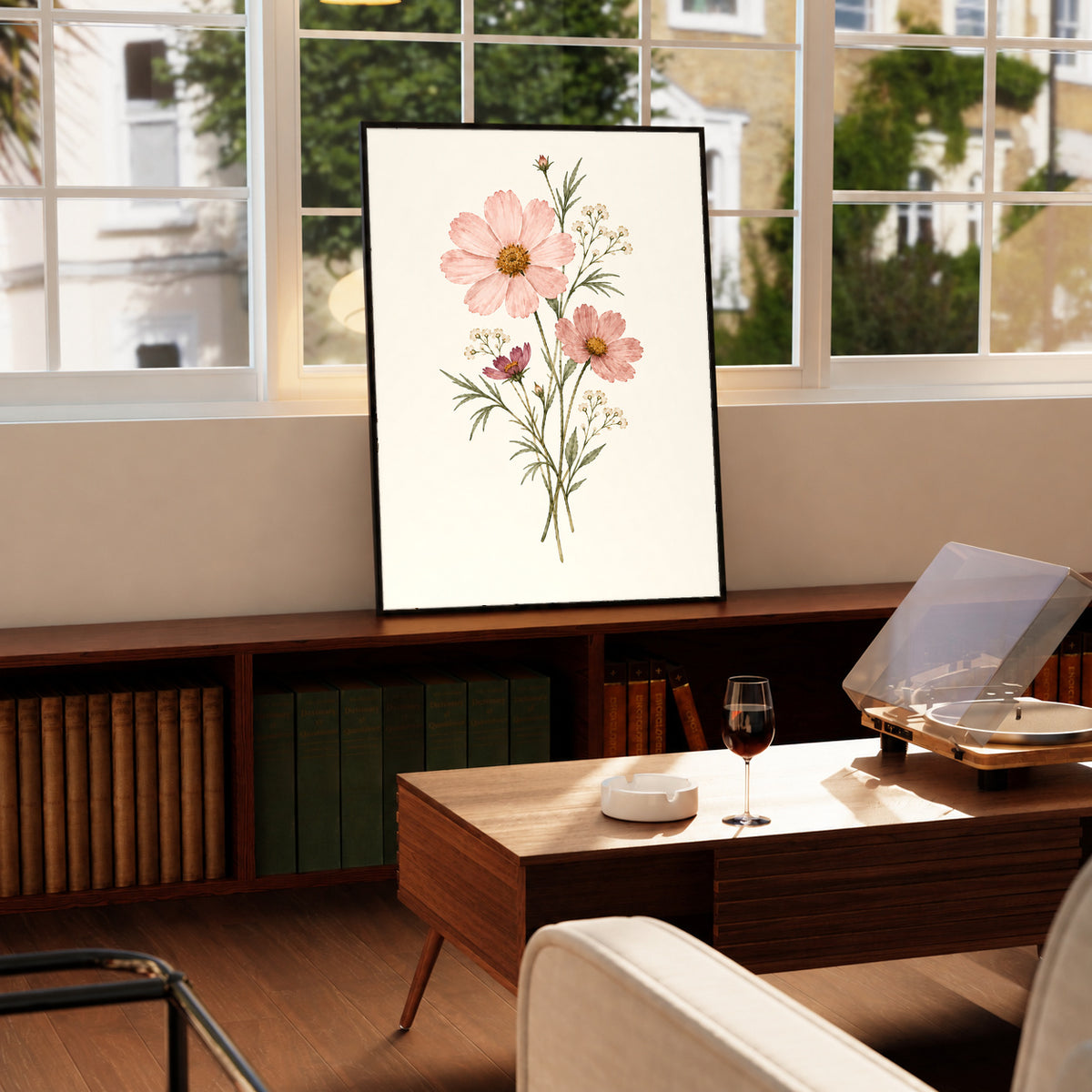 Pink cosmos flowers with yellow centres and fine white botanical sprigs in a black frame on a wooden cabinet beside a window and record player