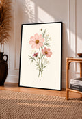 Pink cosmos flowers with yellow centres and fine white botanical sprigs in a black frame leaning on a sunlit wooden floor beside books and a rattan seat