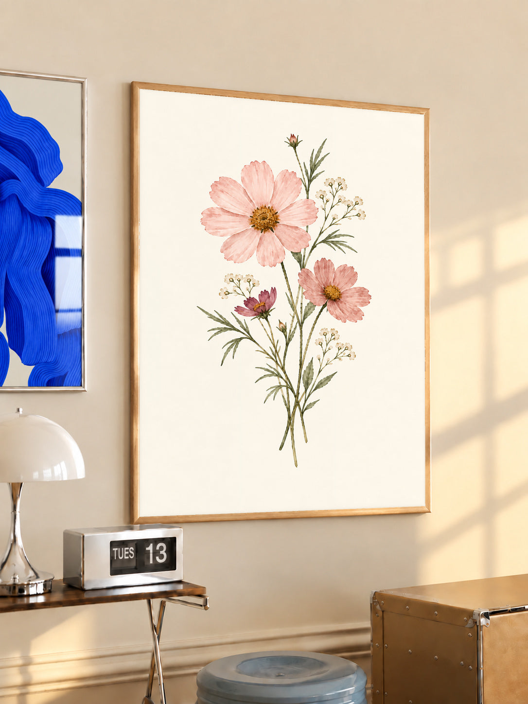 Pink cosmos flowers with yellow centres and fine white botanical sprigs in an oak frame above a console beside a blue print and calendar