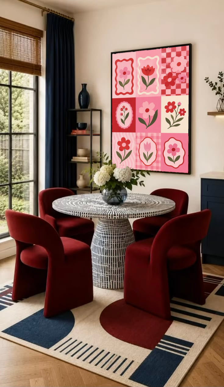 A pink and red patchwork grid of tulips, daisies, scalloped frames and checkerboard squares in a black frame on the wall of a dining nook with red chairs